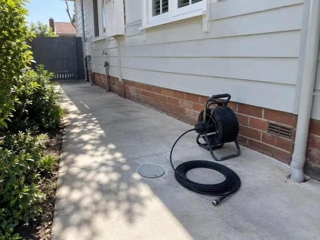 Capped drain inspection opening beside a coiled camera reel on a concrete side path by a house in Mascot