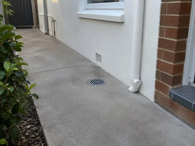 Outdoor gully grate set in dry concrete paving beside a house wall and brick entry in Mascot