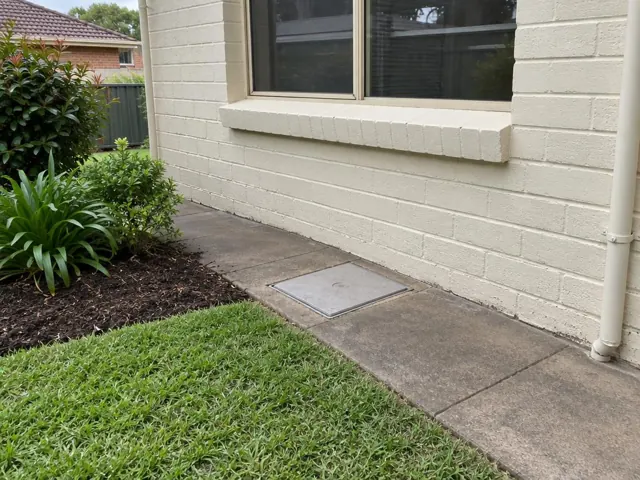 Flush capped access cover in a paved side path beside a cream brick house in Eastlakes