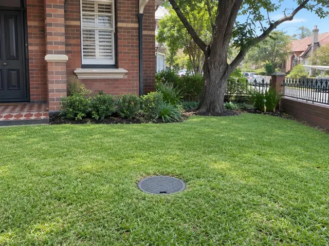 Lawn with a round capped sewer access cover beside a brick house in Botany